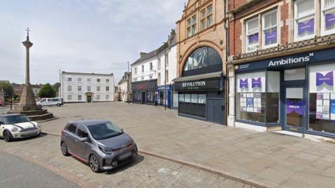 A view of Grantham's Market Place on a sunny day, including a war memorial in the shape of a cross and shopfronts facing the square.