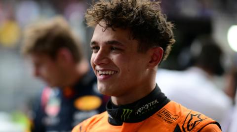 Lando Norris smiling after winning the Sao Paulo Grand Prix, with Max Verstappen visible in the background