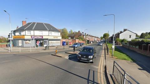 A road junction in a residential area, with a Premier corner shop on the left, grass verges on the right and terrace houses in the middle distance. A black car is waiting at the junction next to a traffic island.