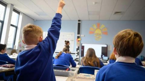 A child in a blue uniform raising his hand in a school classroom. He is sat next to other pupils but we can only see the backs of their heads as they all wear the same uniform of a blue jumper over a white shirt. 