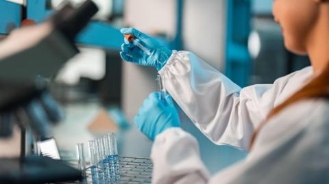 A scientist is is in a lab. They are dressed in a white lab overcoat and are wearing blue medical gloves. They are administering droplets into a test tube. 
Below their hands is a tray of test tubes. 
A microscope is slightly out of focus.