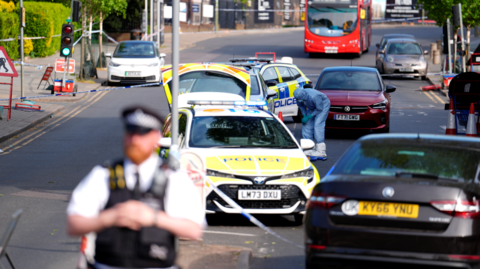 Police cars are parked in the road in the middle of a police cordon around the scene in Golders Green. A scene of crime officer in a blue boiler suit can be seen looking down at the road. A police officer is in the foreground, while an abandoned bus sits in the road in the background.