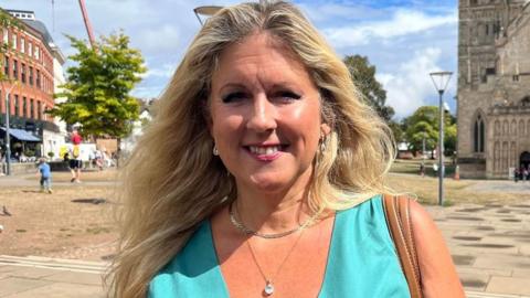 Alison Sheridan, a Conservative member of Exeter City Council, wearing a turquoise dress, a silver necklace and a second necklace with a jewel and the brown straps of a handbag over her shoulder with the front of Exeter Cathedral in the background.