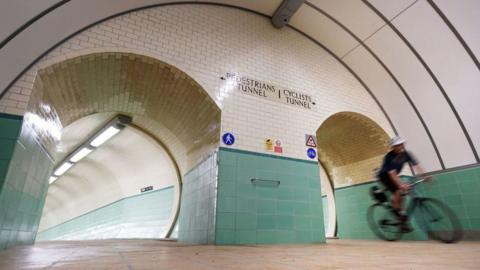 A cyclist rides through the Tyne Pedestrian and Cyclist Tunnel. He is wearing a white helmet and black clothing. The tunnel has blue-green tiles on the lower section and white tiles above. A pedestrian tunnel runs alongside the cycling one.