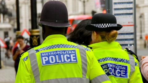 Generic image of two Metropolitan Police officers, wearing high-vis uniform and police hats. The officers, a man and a woman, are pictured from behind outside on a busy London street