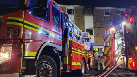 Two fire engines with firefighters seen in between - hoses can be seen coming from the back of one fire engine.  It is night.