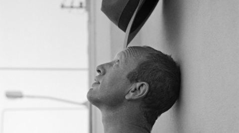 A black and white image of Johnnie Shand Kydd head and neck. It shows his side profile with him leaning his head against a wall while balancing a hat on his head.
