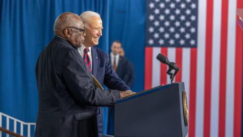 Jim Clyburn appears at a podium with Joe Biden, who is smiling, with an American flag in the background