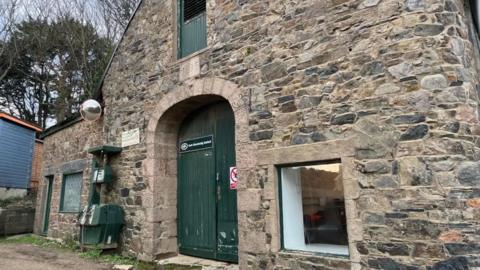 The picture shows an old stone building with green door and window frames. Above the door is a sign that reads 1895. The setting appears rural or wooded, with trees visible in the background.