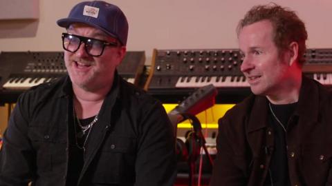 Jez Williams wears black framed glasses and a trilby hat and black jacket and silver necklace, and looks animated as he looks to the left of the camera, sitting down in a studio, and next to him is Andy Williams with short brown hair, with his hands clasped together, wearing a brown jacket, looking over at his brother sitting down 