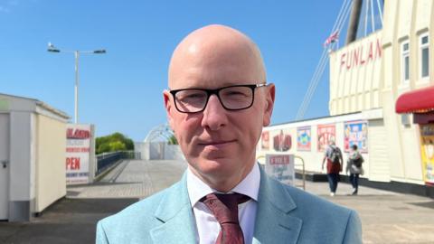 Patrick Hurley wears a pale blue suit, a claret tie and a white shirt. He has dark rimmed glasses and a bald head. Union flags bunting is above his head at the entrance to Southport pier which is partially closed.