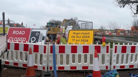 The picture shows road closure red and white stripped fences behind two orange traffic cones. In the background are large metal construction fences and a yellow JCB digger.