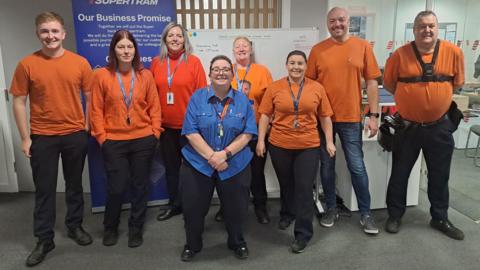 Four men and five women, most wearing orange t-shirts are lined up in an office, smiling  