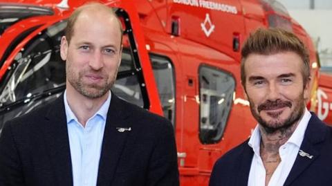 Prince William and David Beckham pose in front of a helicopter