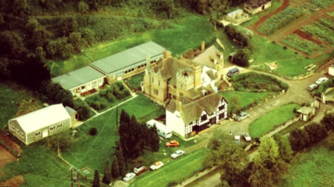 An aerial picture of Berrow Wood School with fields surrounding a black and white Tudor manor. There are cars parked on the road outside and bushes and trees around the edge