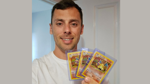 Jon Free, with dark short hair and wearing a white t-shirt, is pictured holding three of his own Pokémon cards while smiling at the camera