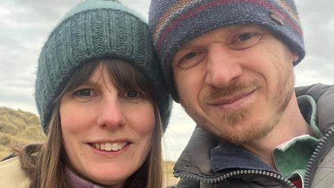 Annette Illing is standing next to her husband Mark on a beach on a cloud day. They are both wearing coats and knitted hats.