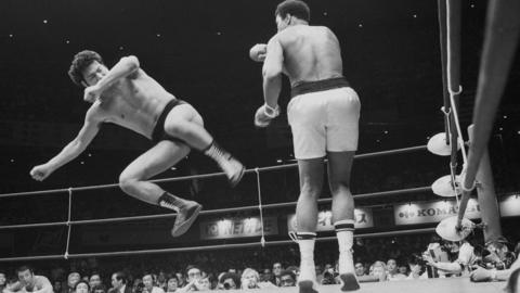 Antonio Inoki is in mid air aiming a kick at Muhammad Ali's legs.