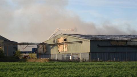 Shot of a large green and grey-coloured industrial shed with plumes of smoke seen rising above it in the background. A fire service aerial ladder is visible in the photograph.