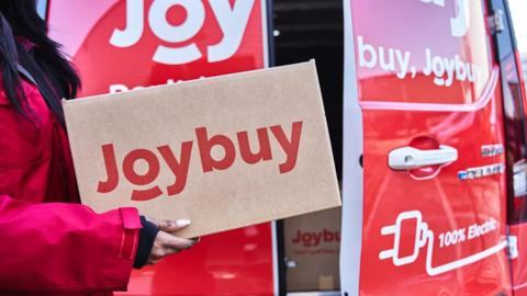 A woman in a red coat is carrying a cardboard box with "Joybuy" printed in red letters. Behind is a red van with a door partially open showing boxes in the back of it.