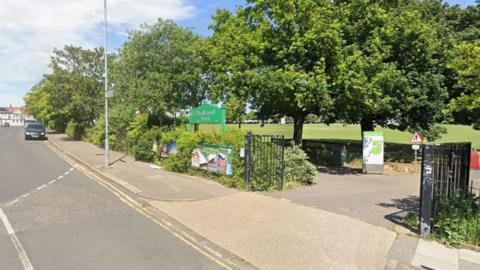 The entrance to Chalkwell Park with open black gates and a green sign with the name on. There are lots of mature trees around the edge, and a large lawn behind them.
