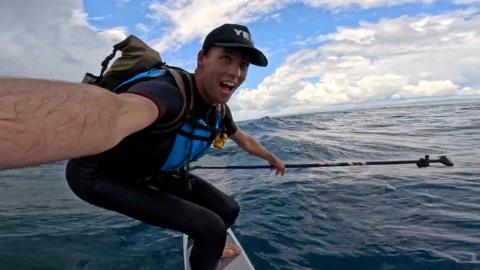A wetsuit‑clad rider balances on a hydrofoil board while holding a paddle, carving over rolling green waves under a bright, cloud-filled sky.