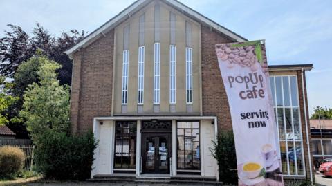 Exterior of a brick Methodist church with long vertical windows above the doorway. Outside the church is a sign advertising a pop up café. 