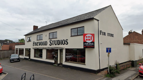 A large white building with six windows at the front and a for sale sign
