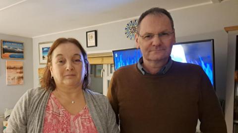 Heather and Nick McLaren look into the camera from their home - she has brown hair, with a pink and white top on and a grey top wrapped around it. He has short dark hair, glasses and a brown jumper on.