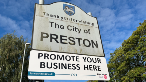 A wide shot of a road sign thanking people for visiting 'the city of Preston'. The white sign is discoloured and dirty.