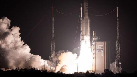 In front of a dark sky, flames and smoke pour from a rocket as it takes off from a launchpad.