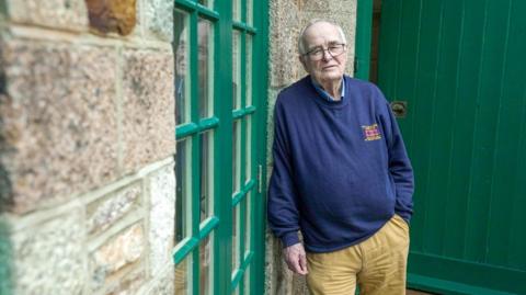 An older man with grey receding hair and glasses leans against a granite wall looking towards the camera. He wears khaki, flannel trousers and a blue sweatshirt. He looks sad.