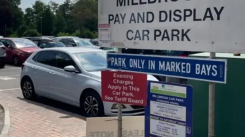 A car park with signage advising of parking costs and to 'park in marked bays'. A white car is the most visible but there are other rows of cars behind. 