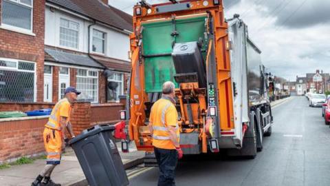 Photo of men in high viz orange work outfits collecting rubbish from wheelie bins and putting it on the back of a bin lorry.