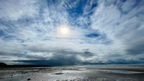 White clouds fill the sky with the sun shining behind in the blue sky and the beach below