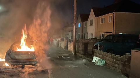 A car which is on fire. It is parked next to a pavement with a row of house on the rigth hand side.