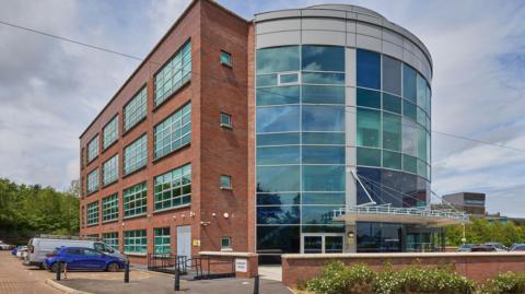 A large four-storey building. One side is mainly red brick with long rectangular windows, the other side mainly glass, with a curved frontage.