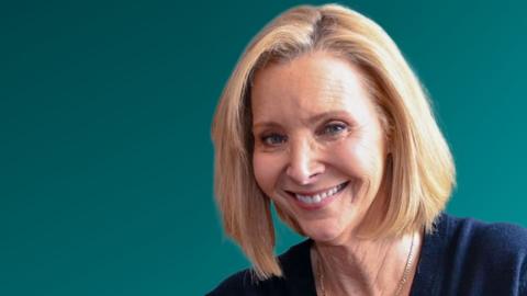 Lisa Kudrow in front of a dark green background. She is wearing a navy blue top.