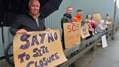 A group of people are standing outside an industrial building in the rain holding handmade signs calling for the site to be saved, including one that reads “SAY NO TO SITE CLOSURE” and another reading “SOS SAVE OUR SITE”.
One person in the foreground is holding an umbrella and a cardboard sign, while others stand along a metal barrier behind them with placards, including a message about saving 167 jobs.