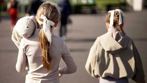 Two girls seen from behind wearing beige and cream clothing with white ribbons in their hair. One is holding a white rabbit soft toy