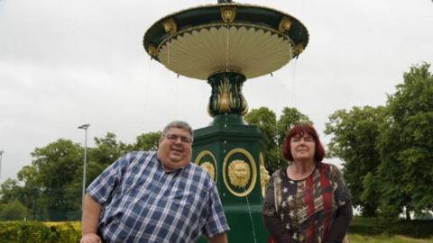 A man in a checked shirt and a woman in a patterned top stand in front of a green-and-gold decorated fountain set in a park.