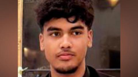 Absi Ahmed looking to the right of the camera lens. He has dark, curly hair which is short on the sides and long on the top. The photo is cropped close, with only his head and shoulders visible. He is wearing a black suit jacket, black shirt and black tie. He has a thin moustache and a beard covering just his lower chin.