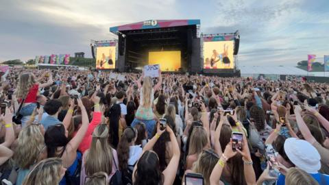 Music fans, some sitting on shoulders with hand-written signs, most filming ton their iphones, crown around the stage in Herrington Country Park. Zara Larsson is seen on large yellow screens wearing a blue dress and being held aloft by her dancers.