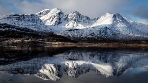 A mountain range covered with snow with the peaks reflected in a loch in the foreground