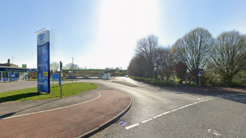 The entrance to a petrol station with trees to the left of the road
