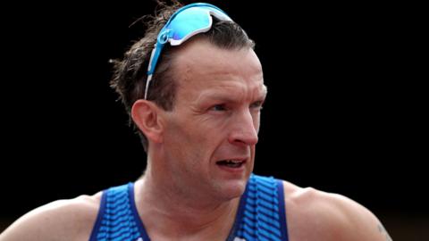 Richard Whitehead, with sunglasses resting on his head. He has dark hair and wears a blue runner vest as he looks to one side.