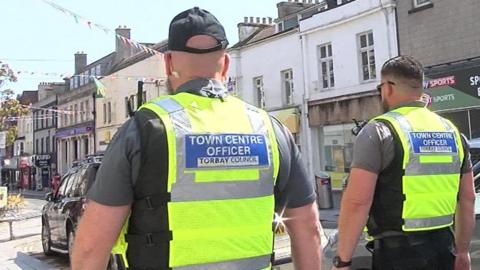 Two Town Centre Officers wearing high visibility clothing. They are walking in a street. There are parked cars and houses with bunting. The sky is blue. 