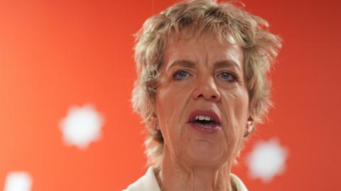 A woman with short blonde hair is pictured talking in front of a red background with white spots. It's a close up but the collar of a white top she is wearing can be seen.