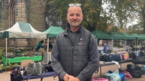 A bald man with glasses resting on his head, and wearing a black jacket and dark grey jumper, stands with his hands clasped in front of him. He is standing in front of a number of market stalls and a number of tables with bags on top of them.