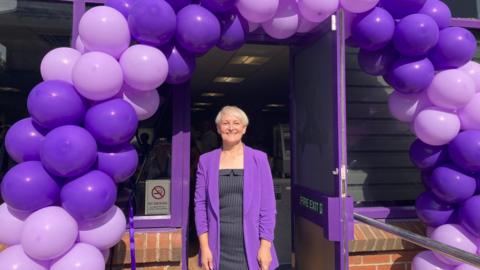 A woman stands at the entrance of a building adorned with an arch of purple and lavender balloons. Dressed in a purple jacket and patterned dress, she faces the open doorway, which leads into a brightly lit interior. A 'No Entry' sign is visible on the adjacent window